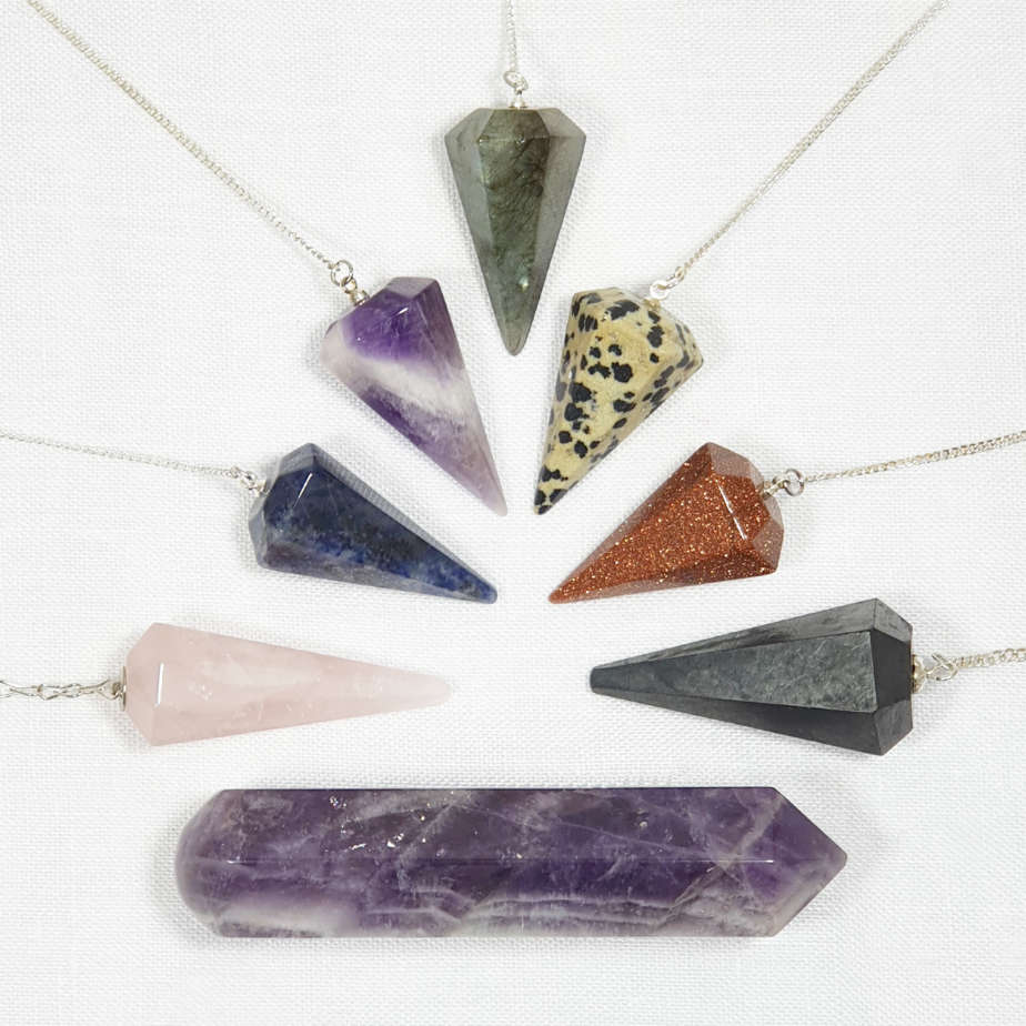 An amethyst wand horizontally positioned above which there are 7 crystal pendulums on silver chains radiating out from a central point